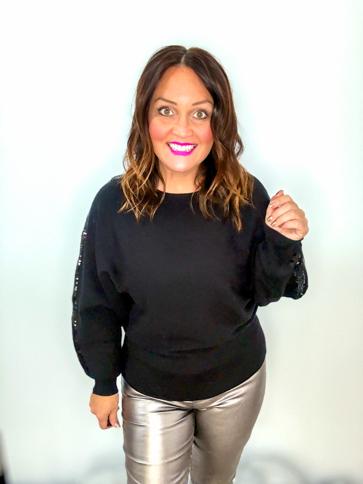 Woman wearing a black glistening sequin sweater, smiling, with shiny pants, showcasing stylish comfort and dazzling design.