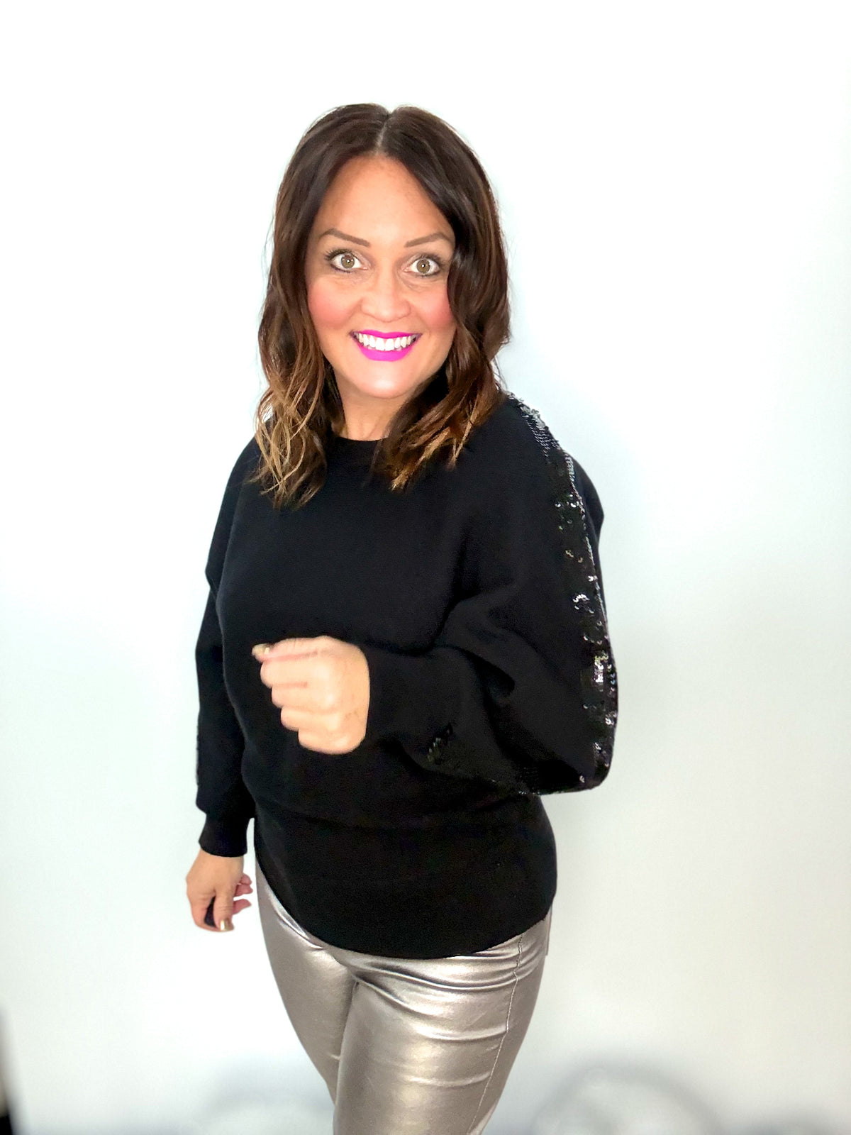 Woman wearing a stylish black sweater with a glistening sequin strip on the sleeve, paired with metallic pants, smiling brightly.