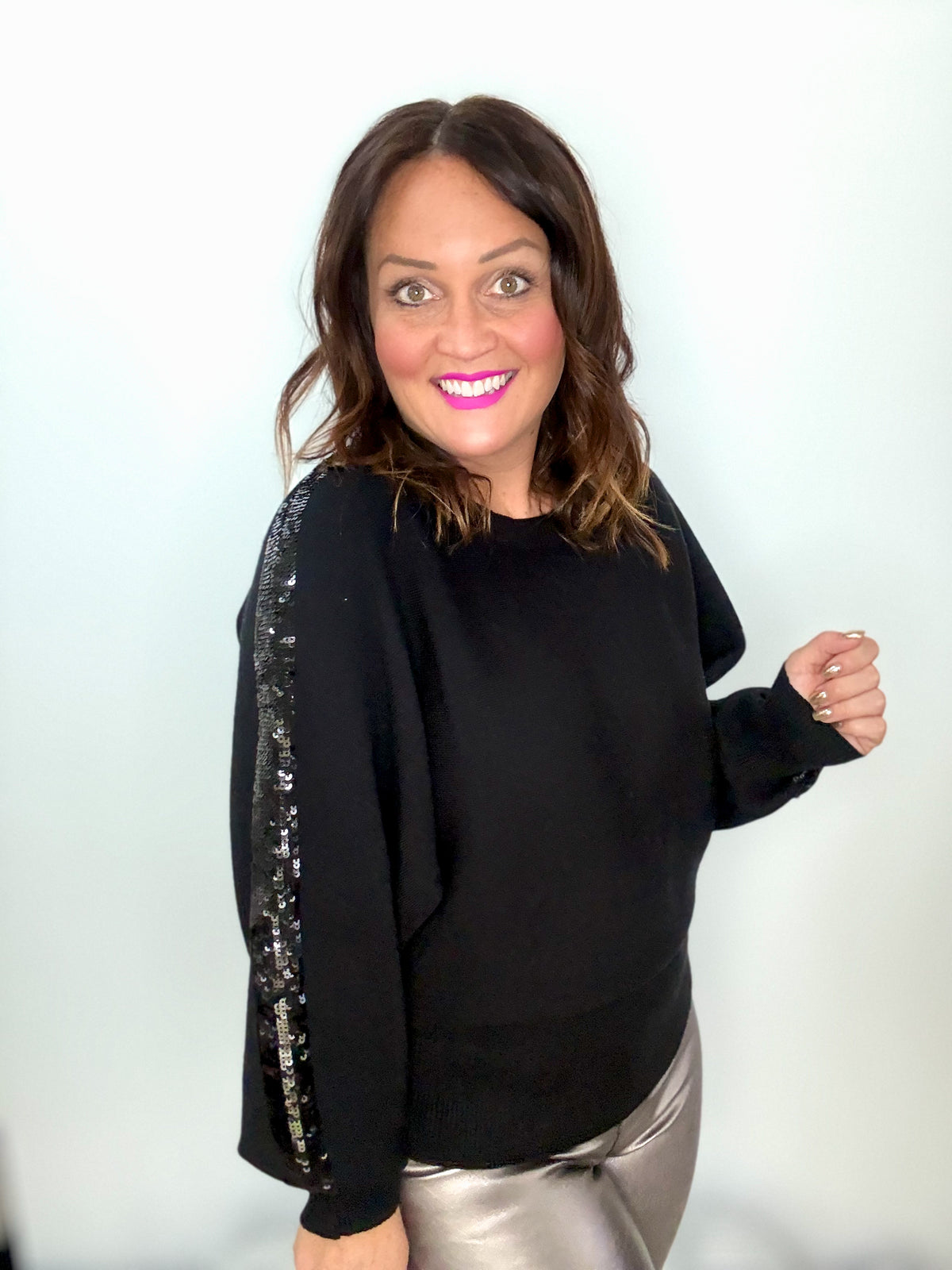 Woman wearing a black sweater with a glistening sequin strip on the sleeve, silver pants, and smiling against a light background.