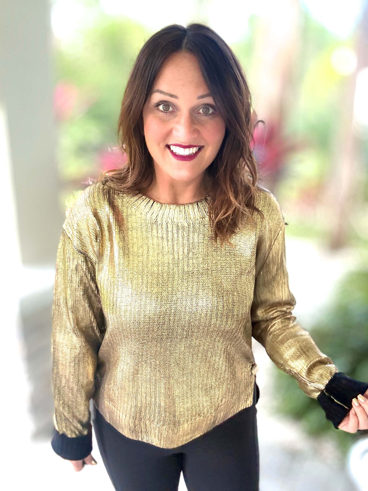 Woman wearing a gold metallic foil sweater with a high low design, featuring black cuffs and a cozy holiday style, smiles outdoors.