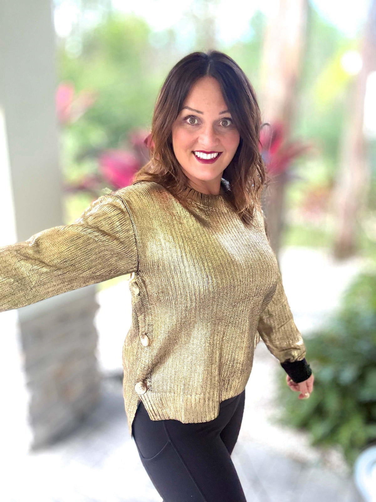 Woman wearing Tis the Season Metallic Foil Sweater in black and gold with button closure, outdoors with greenery in the background.