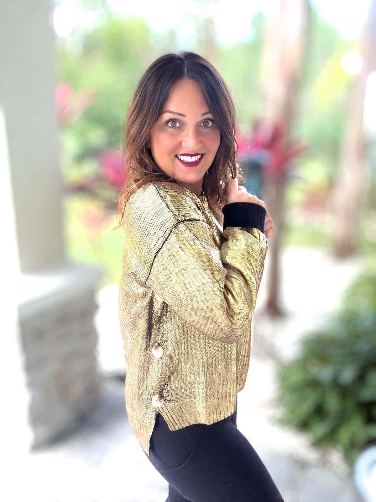 Woman wearing gold metallic foil sweater with button closure, posing outdoors in vibrant garden setting.