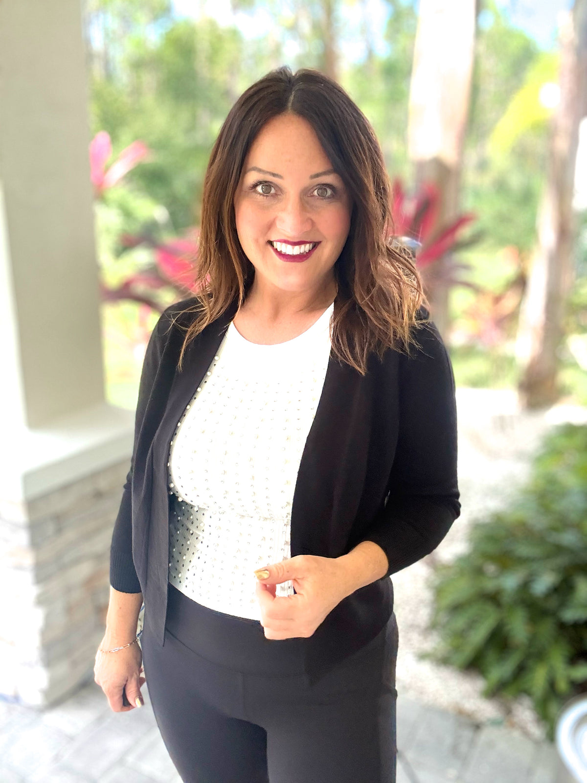 Woman wearing a black knit bolero cardigan, standing outdoors with foliage in the background.