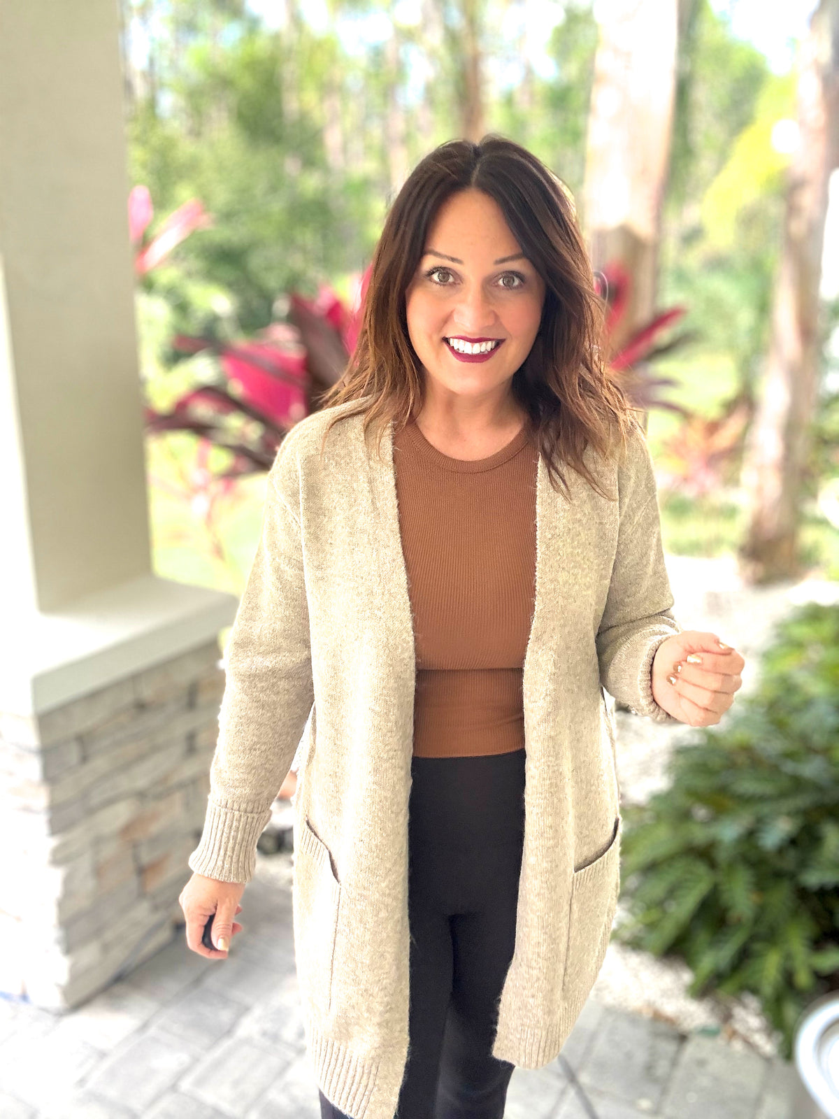 Woman wearing cozy beige cardigan with front pockets, standing outside, surrounded by lush greenery and vibrant plants.